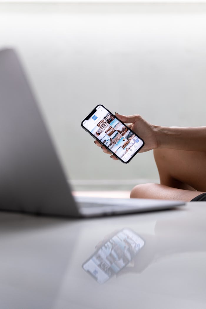 A woman multitasks using a smartphone and laptop for social media in a modern setting.